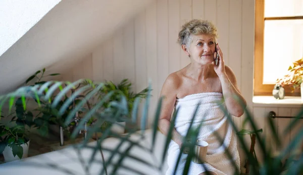 Happy senior woman wrapped in a towel in bathroom at home, resting with tea and smartphone. — Stock Photo, Image