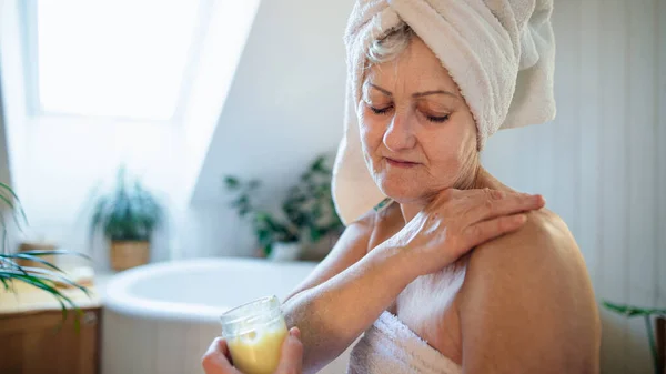 Felice donna anziana avvolta in un asciugamano in bagno a casa, applicando idratante corpo fatto in casa. — Foto Stock