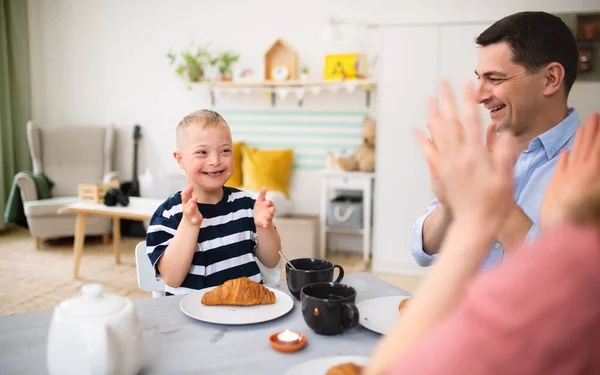 Família feliz com o filho síndrome de down na mesa, batendo palmas ao tomar café da manhã. — Fotografia de Stock