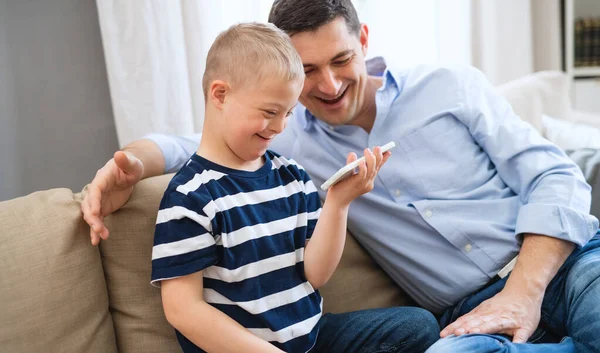 Far med happy down syndrom son inomhus, med hjälp av smartphone för att titta på. — Stockfoto