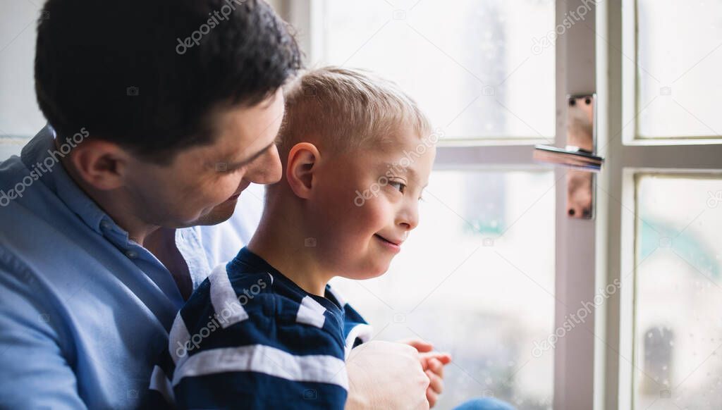 Father with happy down syndrome son indoors at home, looking through window.