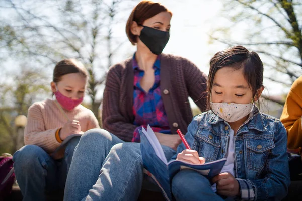 Вчителька з маленькими дітьми, сидячи на відкритому повітрі в міському парку, навчаючись груповій освіті та концепції коронавірусу . — стокове фото