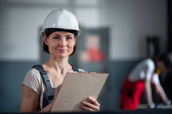 Portret van een vrouw werknemer met helm binnen in de fabriek houden klembord. — Stockfoto