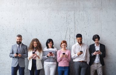 Ofisin içinde beton duvara karşı duran, akıllı telefon kullanan girişimci grubunun portresi.
