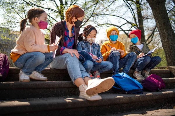 Lärare med små barn som sitter utomhus i stadsparken, lärande grupp utbildning och coronavirus koncept. — Stockfoto