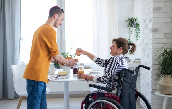 Disabled mature woman in wheelchair sitting at the table with a son indoors at home, eating. — Stock Photo, Image