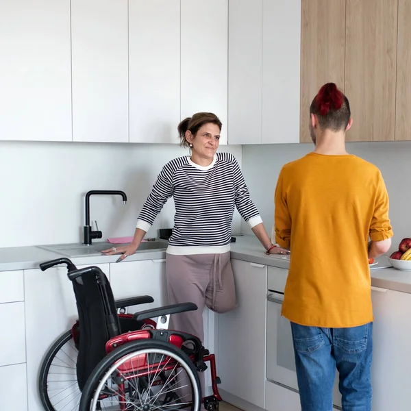 Disabled mature woman in wheelchair talking to son in kitchen indoors at home. — Stock Photo, Image