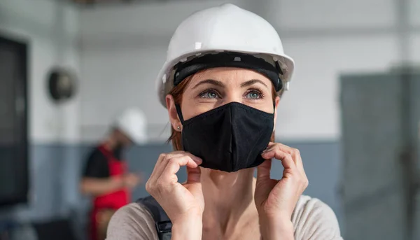 Portrait of woman worker with helmet indoors in factory putting on face mask. — Stock Photo, Image