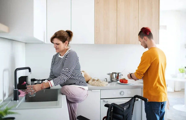 Funktionshindrad mogen kvinna i rullstol pratar med son i köket inomhus. — Stockfoto