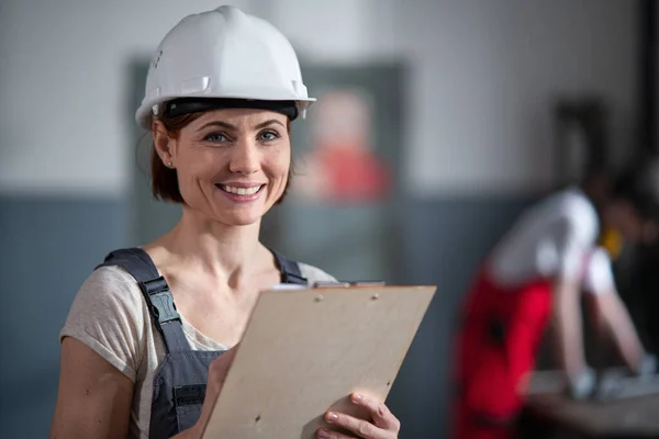 Retrato de mulher trabalhador com capacete dentro de casa na fábrica segurando prancheta. — Fotografia de Stock