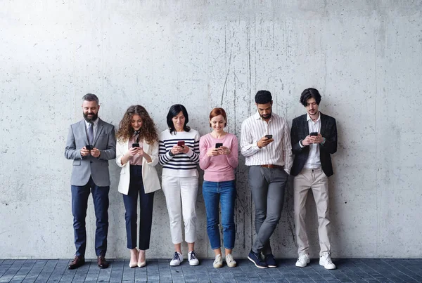 Portrét skupiny podnikatelů stojících u betonové zdi v kanceláři, pomocí smartphonu. — Stock fotografie