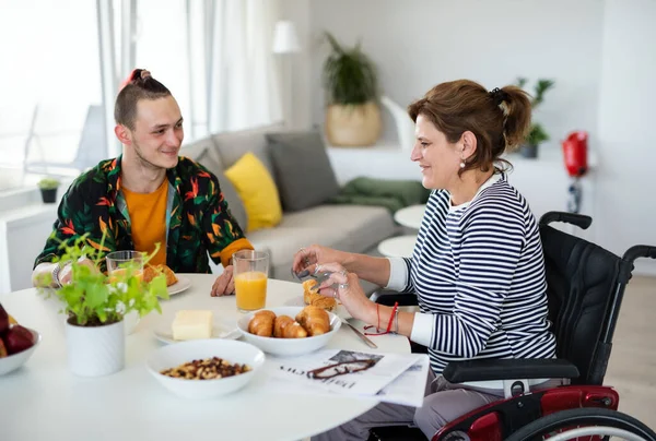 Αναπηρία ώριμη γυναίκα σε αναπηρική καρέκλα κάθεται στο τραπέζι με ένα γιο σε εσωτερικούς χώρους στο σπίτι, τρώει. — Φωτογραφία Αρχείου