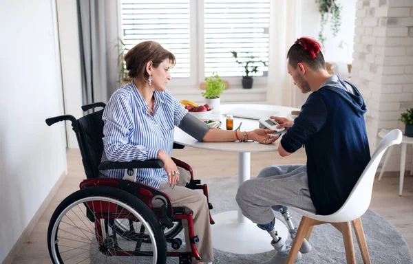Zdravotně postižení lidé na invalidním vozíku sedí doma u stolu, měří krevní tlak. — Stock fotografie