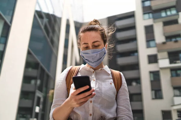 Портрет молодой женщины в маске с использованием смартфона на открытом воздухе в городе, жизнь после вакцинации ковид-19. — стоковое фото