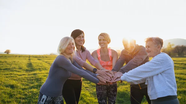 Ομάδα ηλικιωμένων με τον δάσκαλο του αθλητισμού στοίβαγμα χέρια μαζί μετά την άσκηση σε εξωτερικούς χώρους στη φύση κατά το ηλιοβασίλεμα. — Φωτογραφία Αρχείου