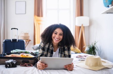Young woman with tablet and suitcase packing for holiday at home, looking at camera.