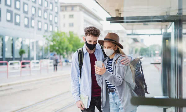 Молодая пара туристических путешественников со смартфоном на отдыхе на автобусной остановке в городе, коронавирус концепции. — стоковое фото