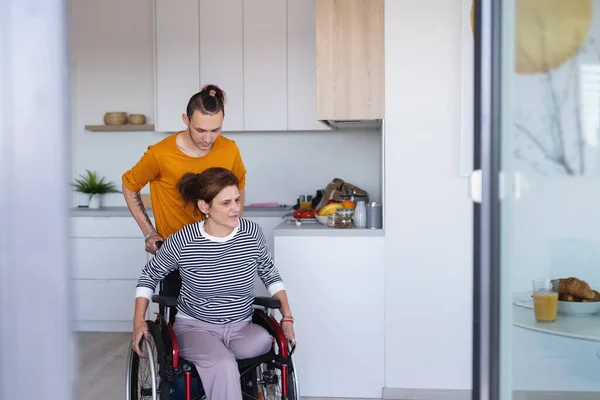 Adult son helping disabled mature mother in wheelchair in kitchen indoors at home. — Stock Photo, Image