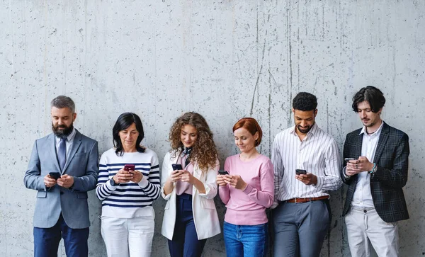 Portret van een groep ondernemers die binnen op kantoor tegen een betonnen muur staan, met smartphone. — Stockfoto