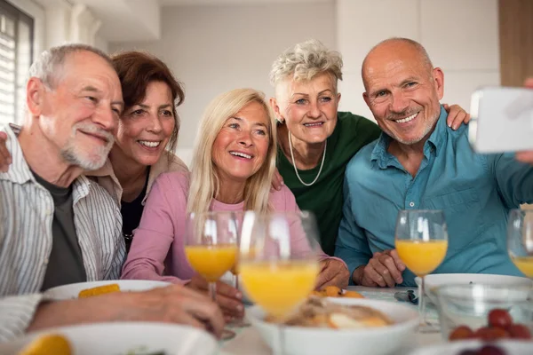 Группа старших друзей устраивает вечеринку в помещении, делает селфи во время еды за столом. — стоковое фото