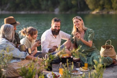 Yaz tatili gezisinde mutlu çok nesilli aile, göl kenarında barbekü.