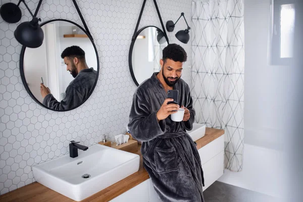 Jeune homme avec café et peignoir à l'intérieur dans la salle de bain à la maison, prendre selfie. — Photo