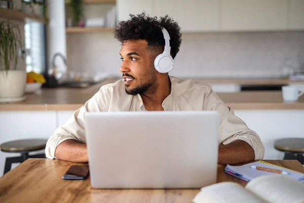 Young man student with laptop and headphones indoors at home, studying. — Stock Photo, Image