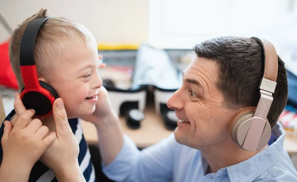 Vader met happy down syndroom zoon binnen thuis, met behulp van een koptelefoon. — Stockfoto