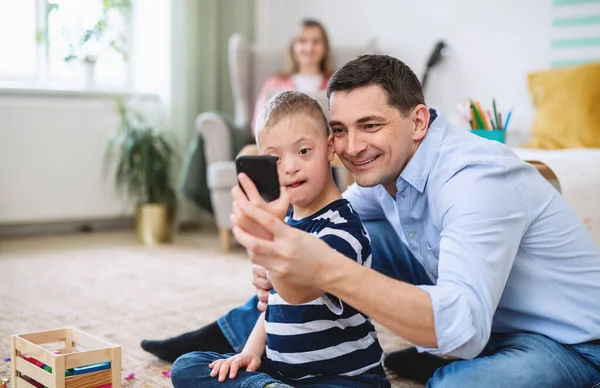 Far med happy down syndrom son inomhus hemma, tar selfie med smartphone. — Stockfoto