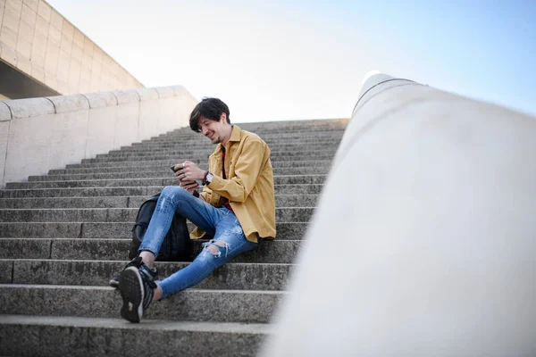 Portrét mladého muže sedícího venku na schodišti ve městě, používajícího smartphone. — Stock fotografie