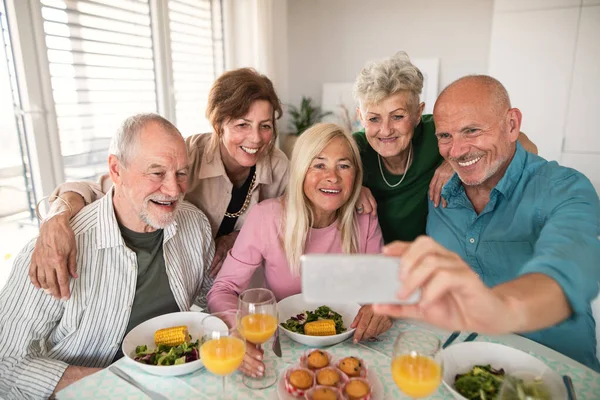 Gruppe älterer Freunde feiert drinnen, macht Selfie beim Essen am Tisch. — Stockfoto