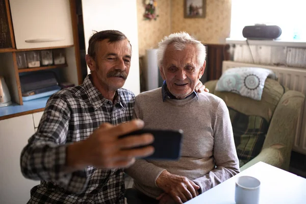 Portret mężczyzny ze starszym ojcem siedzącym przy stole w domu, robiącym selfie. — Zdjęcie stockowe