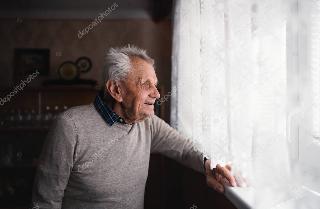 Portrait of elderly man standing indoors at home, looking out through window.
