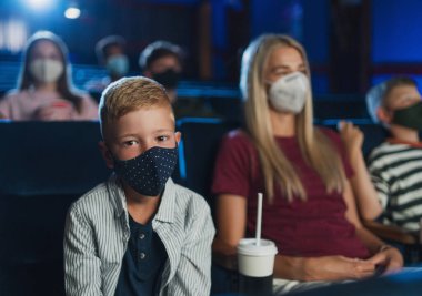 Annesi olan küçük bir çocuk sinemada kameraya bakıyor, film izliyor ve koronavirüs konsepti uyguluyor..