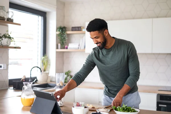 Jovem com tablet preparando café da manhã saudável em casa, conceito de escritório em casa. — Fotografia de Stock