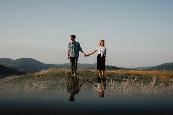 Pareja joven de pie en la naturaleza en el campo, tomados de la mano, pero mirando el uno al otro. —  Fotos de Stock