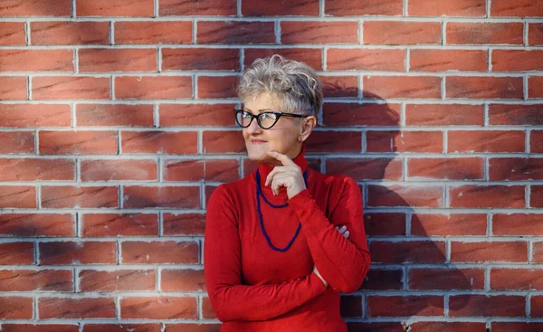 Senior woman standing outdoors against brick wall background. Copy space. — Stock Photo, Image