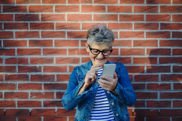 Старшая женщина стоит на улице на фоне кирпичной стены, используя смартфон. — стоковое фото