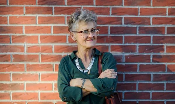 Senior woman standing outdoors against brick wall in town, arms crossed. — Stock Photo, Image