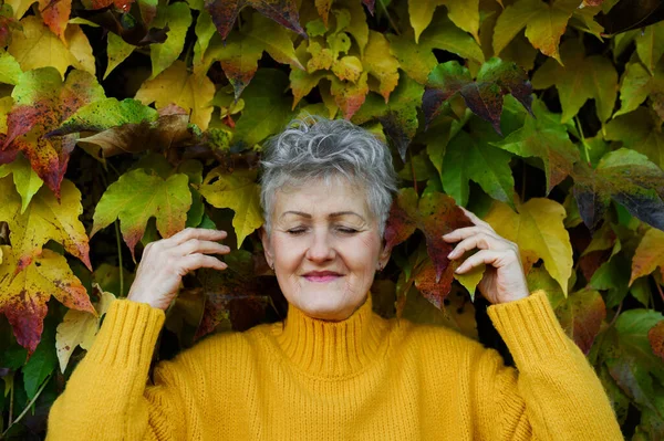 Mujer mayor de pie al aire libre contra el colorido fondo natural de otoño, los ojos cerrados. — Foto de Stock