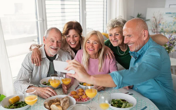 Gruppe älterer Freunde feiert drinnen, macht Selfie beim Essen am Tisch. — Stockfoto