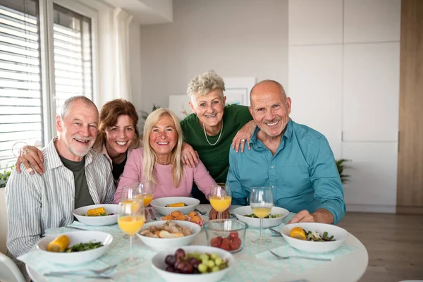 Gruppo di amici anziani che fanno festa in casa, guardando la fotocamera quando si mangia. — Foto Stock