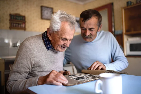 Portret mężczyzny ze starszym ojcem siedzącym przy stole w domu, patrzącym na album fotograficzny. — Zdjęcie stockowe