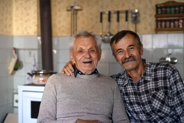 Portret mężczyzny ze starszym ojcem siedzącym przy stole w domu, patrzącym w kamerę. — Zdjęcie stockowe