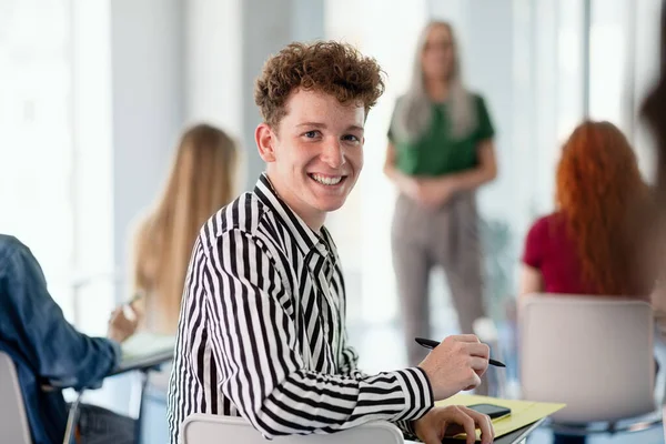 Portret van een jonge universiteitsstudent die binnen in de klas zit en naar de camera kijkt. — Stockfoto