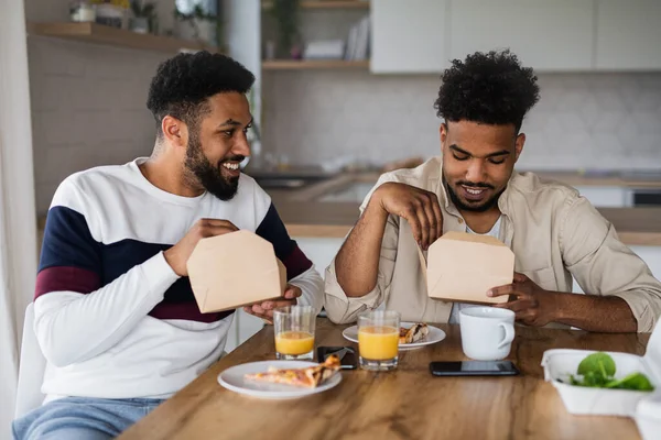 Porträt junger erwachsener Brüder, die zu Hause in der Küche sitzen und Essen zum Mitnehmen essen. — Stockfoto