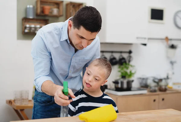 Pai com feliz síndrome de down filho dentro de casa na cozinha, mesa de limpeza. — Fotografia de Stock