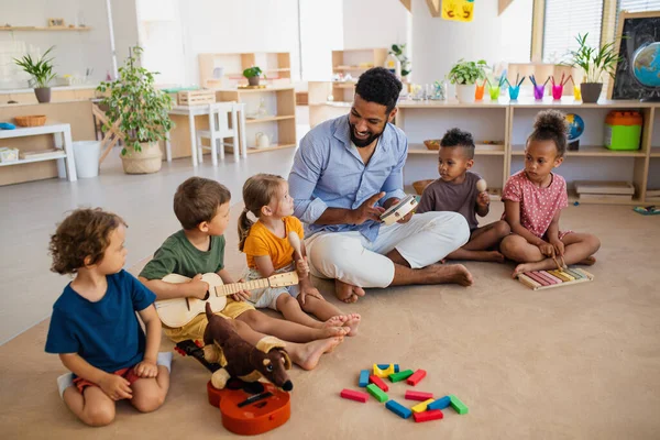 Gruppe kleiner Kindergartenkinder mit einem männlichen Lehrer, der im Klassenzimmer auf dem Boden sitzt und Musikinstrumente spielt. — Stockfoto