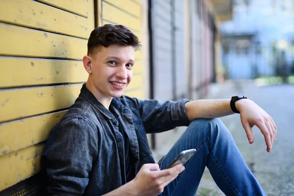 Portrait of young man with smartphone sitting outdoors in city street, looking at camera. — Stock Photo, Image