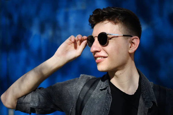 Portrait of young man with sunglasses standing outdoors against blue background. — Stock Photo, Image